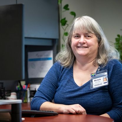 Michelle Hiebert is pictured sitting at her desk, located at the HDH site. She has shoulder-length, silver hair and is wearing a blue, long-sleeved shirt.