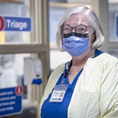 Nancey Martindale pictured inside KHSC's Emergency Department has short grey hair. She wear glasses and is photographed wearing a mask, blue scrubs and a yellow gown.