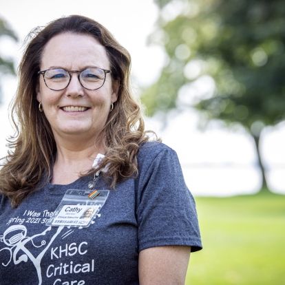 Cathy Seymour has long brown, wavy hair. She has glasses and is wearing a grey t-shirt. She's pictured standing outside the Kingston General Hospital site.