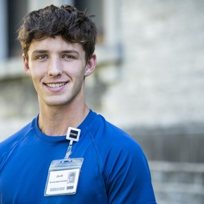 Jacob Kasaboski has short, dark, curly hair. He's wearing a bright, blue top and standing outside the Kingston General Hospital site.