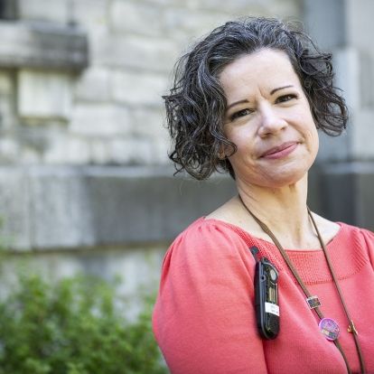 Jane Lewis has short, dark and wavy hair, and dark brown eyes. She’s wearing a bright coral blouse and is pictured standing outside Kingston General Hospital.