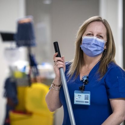 Kimberley Dickinson is pictured holding a broom in a hallway at the HDH site. She has blonde, shoulder length hair and is wearing blue scrubs and mask.