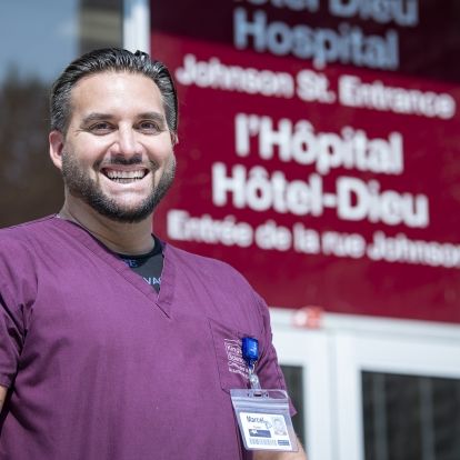 Marcel Cernik has short dark hair and a beard. He's wearing purple scrubs and pictured standing outside of Hotel Dieu Hospital.