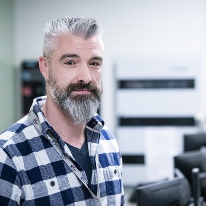 Mark Westcott is pictured in the Switchboard department at the KGH site. He has dark brown eyes, salt and pepper, spiky hair and a beard. He’s wearing a blue and white plaid, long-sleeved shirt.