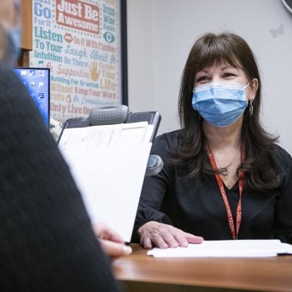 Sandra Mahoney is pictured sitting at her desk in Johnson 5 at the HDH site. She has dark brown, shoulder length hair and brown eyes. She’s wearing a black blouse and mask.