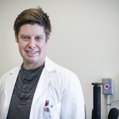 Tim Woodford is pictured inside KHSC’s electroencephalography (EEG) services department. He’s wearing a white lab coat with a grey shirt underneath. He has blue eyes and short, light brown hair.