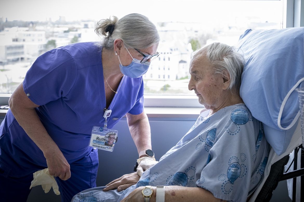 PoKHSC - Ruth Bradshaw Ruth Bradshaw is pictured caring for a patient in a wheelchair in their room. Bradshaw has white/silver hair, pulled back into a bun, black framed glasses and is wearing blue scrubs with a mask. She’s leaning over the patient and her hand is on his arm in a loving way.