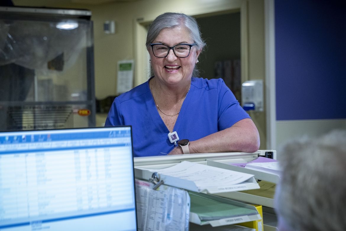 PoKHSC - Ruth Bradshaw Ruth Bradshaw is pictured smiling and chatting with a colleague, while standing behind a care desk at Kingston General Hospital. She has white/silver hair, pulled back into a bun, black framed glasses and is wearing blue scrubs.