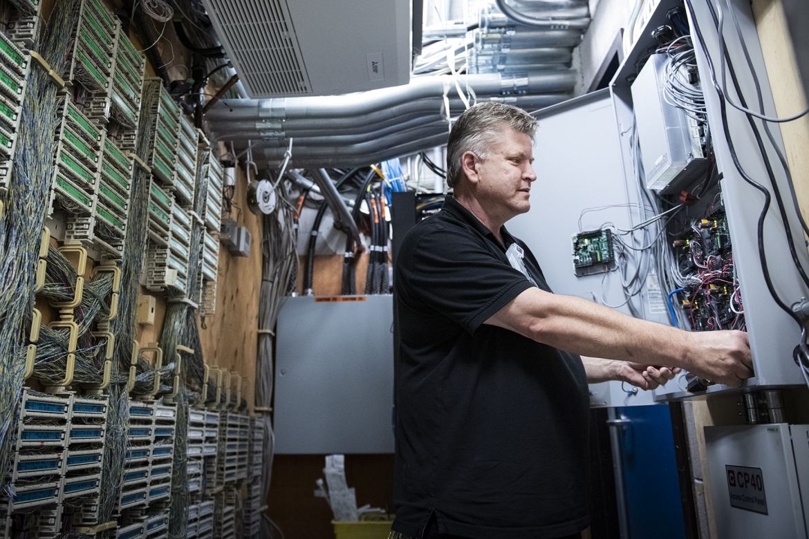 People of KHSC - Andrew Schick Andrew Schick is photographed working on equipment at the Kingston General Hospital site. He has short, salt and pepper hair, blue eyes and is wearing a black polo shirt.