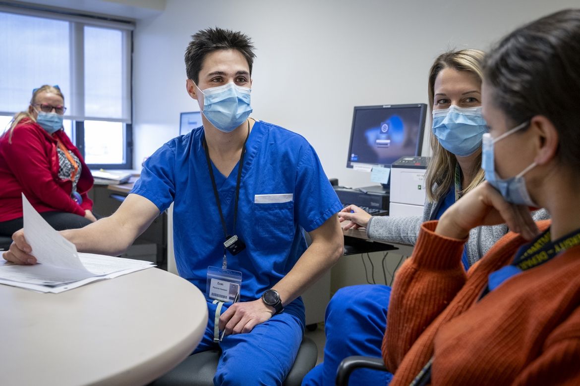 People of KHSC: Evan Earl Evan Earl is seated next to two women around a circular table, another woman is seen seating in the background. Earl has short, dark, spiky hair, brown eyes, and is wearing a mask and bright blue scrubs. The two woman are also wearing masks and bright blue scrubs.