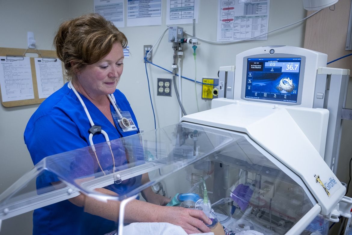 People of KHSC - Julie Vanyolai Julie Vanyolai is pictured caring for a young patient in an incubator/isolette. She has strawberry blonde/red hair, which is tied up in a bun, and blue eyes. She’s wearing blue scrubs and has a stethoscope around her neck.