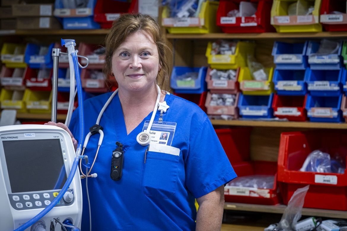 People of KHSC - Julie Vanyolai Julie Vanyolai is pictured standing next to a piece of respiratory therapy equipment, and is looking directly into the camera. She has strawberry blonde/red hair, which is tied up in a bun, and blue eyes. She’s wearing blue scrubs and has a stethoscope around her neck.