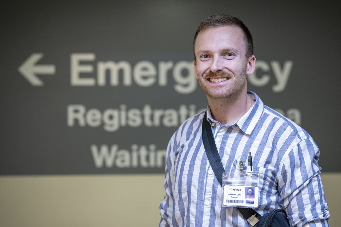 PeopleOfKHSC_Khayman_Wood_2 Khayman Wood is wearing a striped shirt and his KHSC ID badge, while standing smiling directly to the camera in a hallway outside the Emergency Department. The sign in the background has the words "emergency," "registration area," and "waiting" partially visible, with an arrow pointing to the left.
