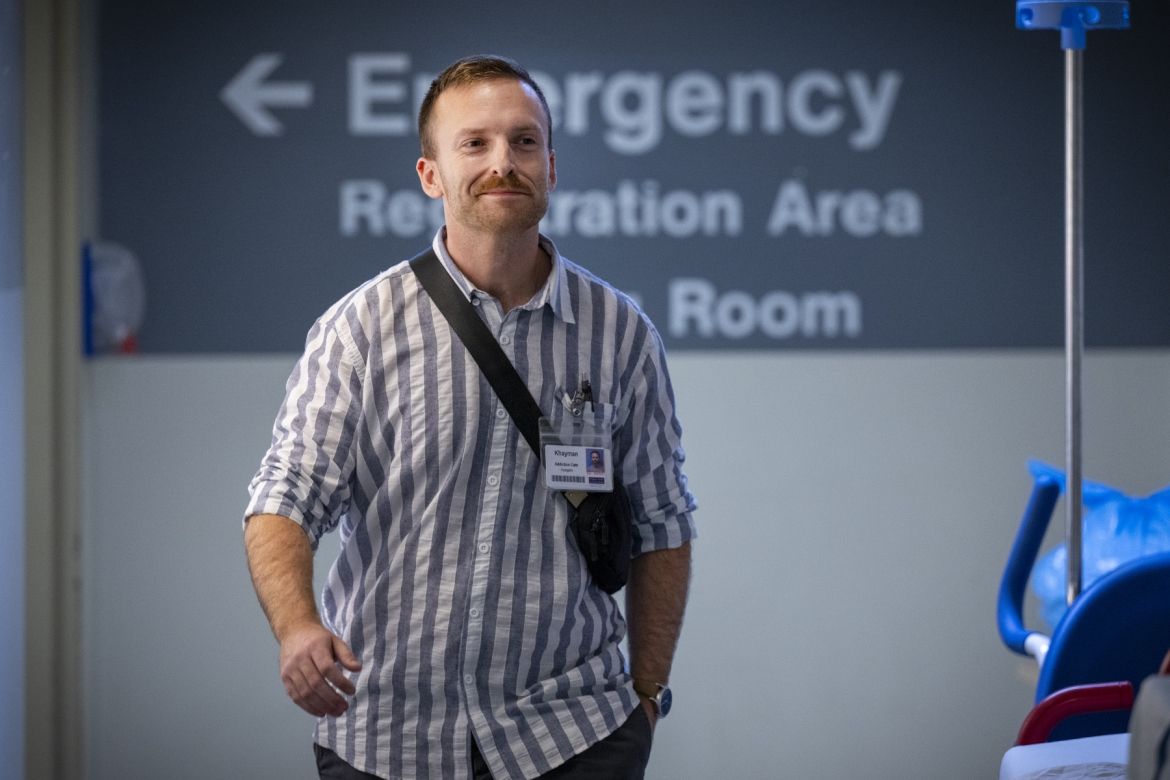 PeopleOfKHSC_Khayman_Wood_3 Khayman Wood is walking down a hallway at the Kingston General Hospital site. Behind him, a sign reads "emergency registration area room" with an arrow pointing to the left. A blue medical chair and equipment are partially visible on the right side of the image.