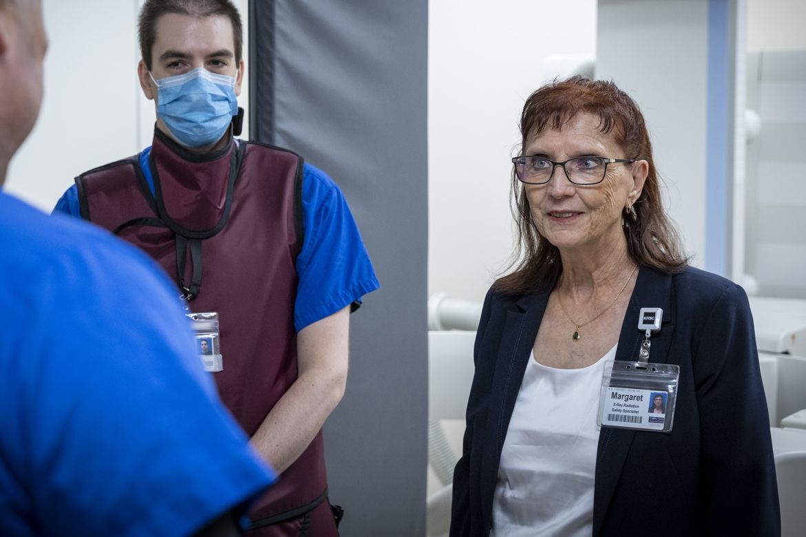 People of KHSC - Margaret Bollen Margaret Bollen is pictured standing in a suite at Kingston General Hospital. She has auburn hair, blue eye hair and wears glasses. She’s wearing a dark navy suit and white top. She’s standing next to a medical radiation technologist.