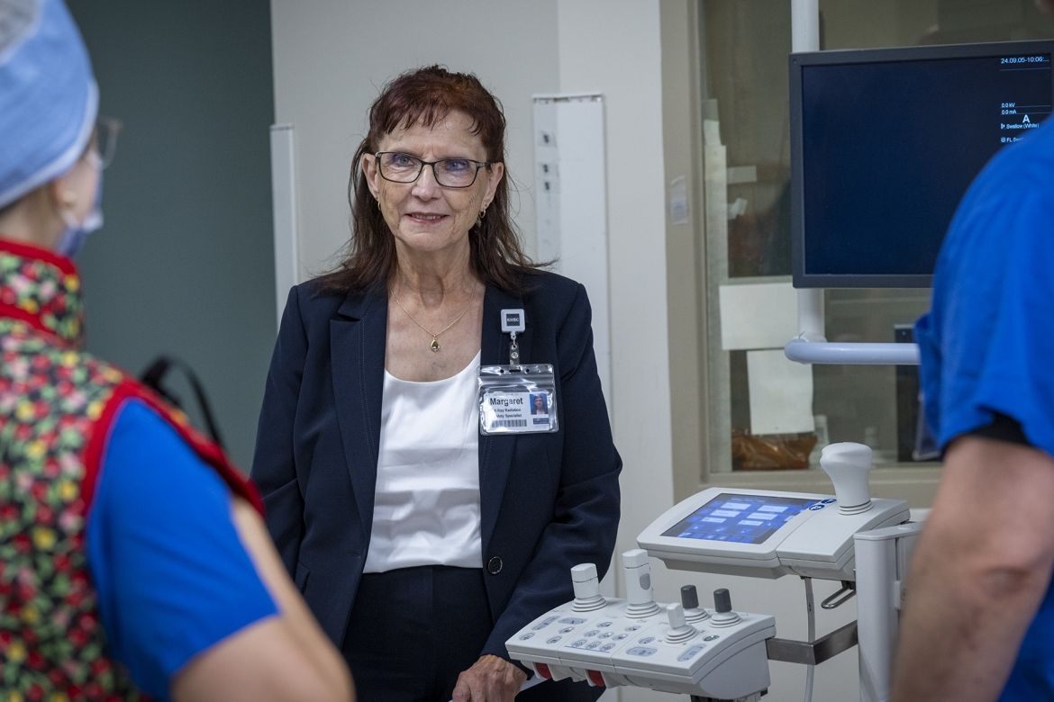 People of KHSC - Margaret Bollen Margaret Bollen is pictured standing in a suite at Kingston General Hospital, next to a piece of equipment. She has auburn hair, blue eye hair and wears glasses. She’s wearing a dark navy suit and white top. She’s chatting with a medical radiation technologist.