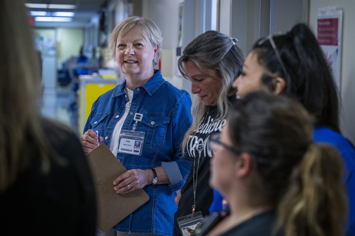 People of KHSC - Patti Cox Patti Cox is pictured taking part of team huddle on a patient care unit. She’s smiling as a member of the care team speaks to the group. Cox has blue eyes and light blonde hair styled in a pixie cut. She’s wearing a blue denim blouse on top of a white t-shirt, and is holding a clipboard and pen.