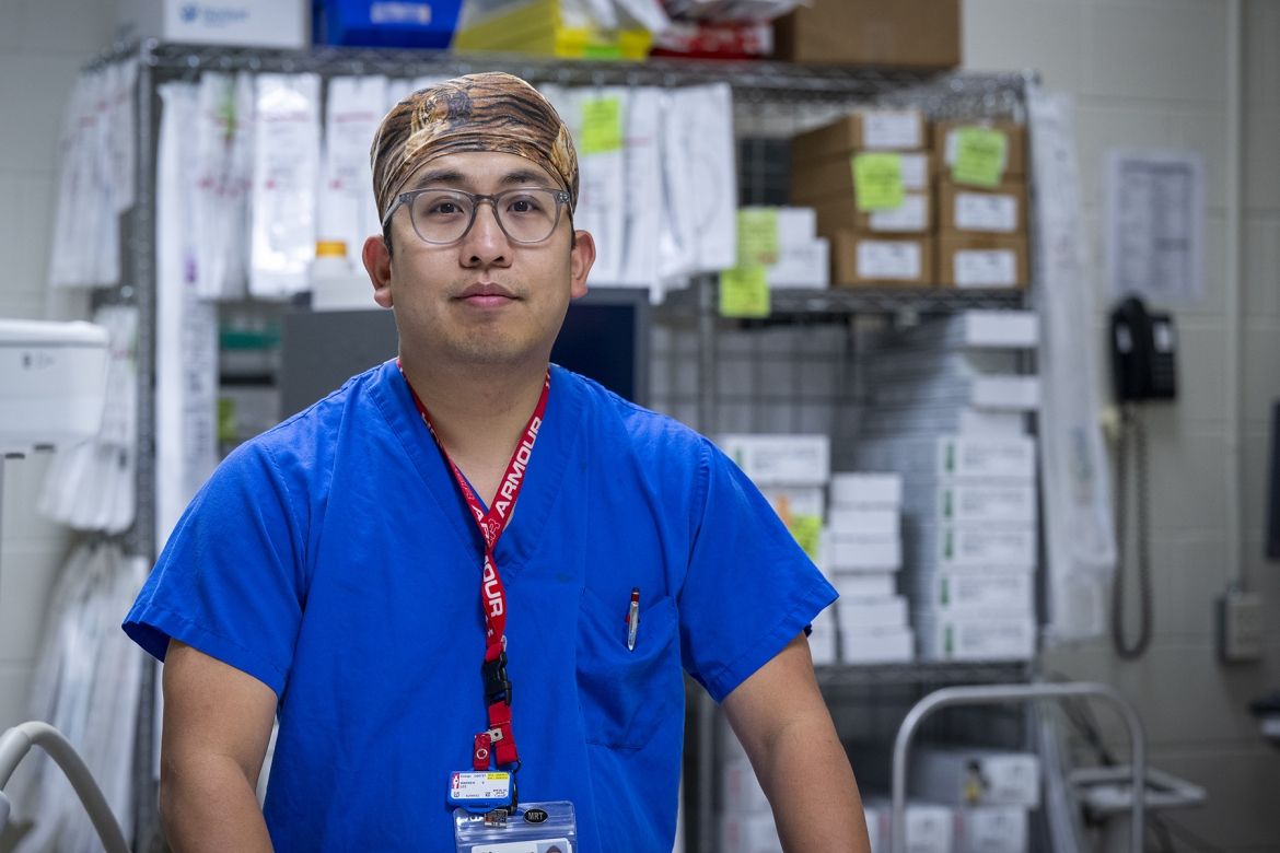PeopleOfKHSC_Warren_Lee_1 Warren Lee is pictured standing in room filled with medical equipment at the Kingston General Hospital site. He’s facing the camera directly and wearing bright, blue scrubs, a tiger print scrub cap, and has grey, translucent glasses.