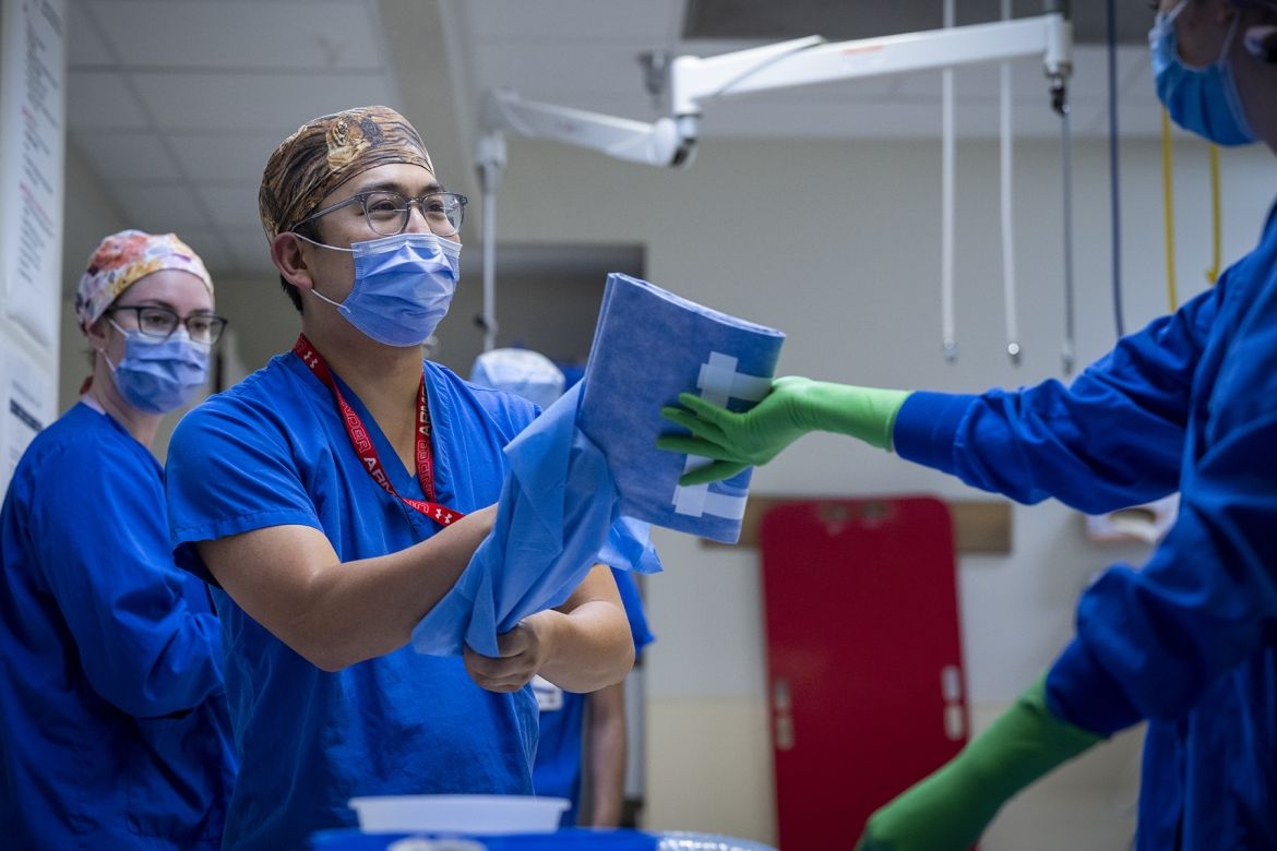 PeopleOfKHSC_Warren_Lee_2 Warren Lee is pictured in a room with two other colleagues, who are getting ready for a procedure. Lee is wearing bright, blue scrubs, tiger print scrub cap, a mask over his nose and mouth, and has grey, translucent glasses. He’s facing a colleague who is handing him some blue clothes.