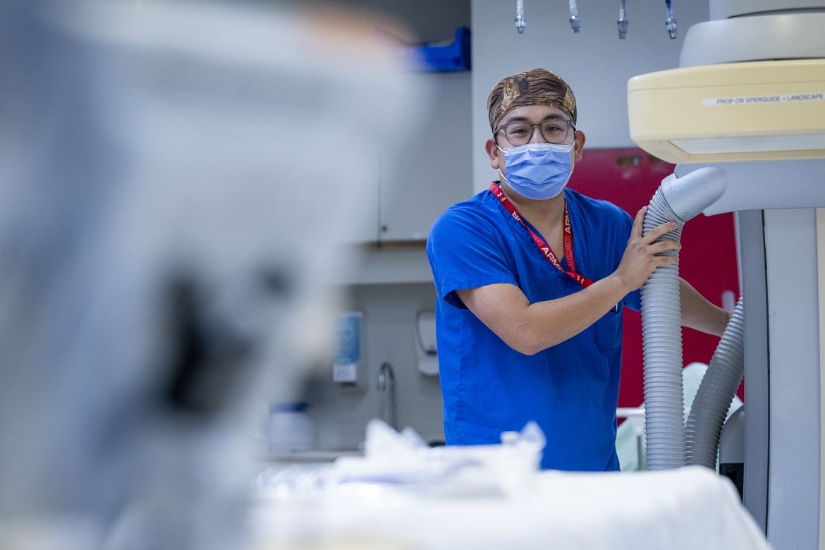 PeopleOfKHSC_Warren_Lee_4 Warren Lee is pictured in the background of an image next to a piece of equipment. He’s holding the equipment while looking directly at the camera. Lee is wearing bright, blue scrubs, tiger print scrub cap, a mask over his nose and mouth, and has grey, translucent glasses.