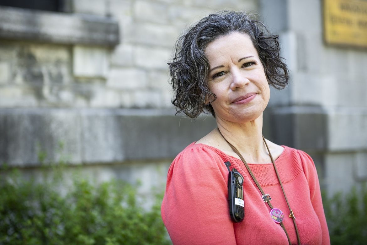 Jane Lewis has short, dark and wavy hair, and dark brown eyes. She’s wearing a bright coral blouse and is pictured standing outside Kingston General Hospital.