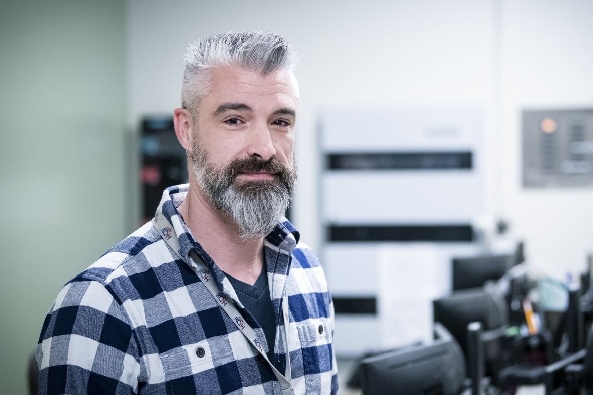 Mark Westcott is pictured in the Switchboard department at the KGH site. He has dark brown eyes, salt and pepper, spiky hair and a beard. He’s wearing a blue and white plaid, long-sleeved shirt.
