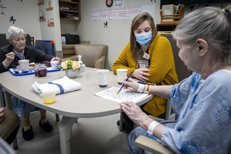 Jessica McCutcheon is seated in between two elderly female patients. One patient is doing arts and crafts, while the other eats a meal. McCutcheon has dark blonde hair and brown eyes. She’s wearing a black top, mustard cardigan and a mask over her mouth and nose.