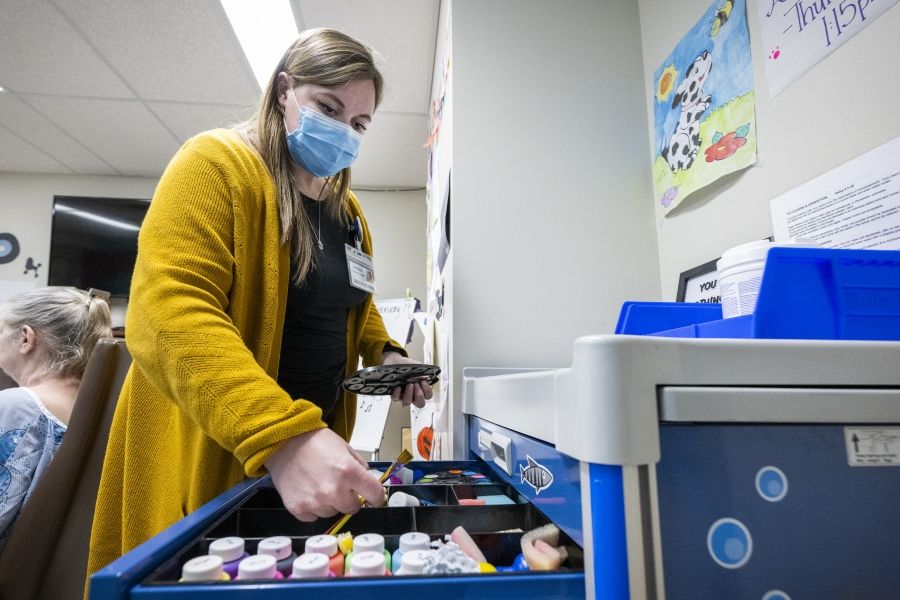 Jessica McCutcheon is standing in front of a drawer, gathering paint, brushes and other art colourful art supplies. McCutcheon has dark blonde hair and brown eyes. She’s wearing a black top, mustard cardigan and a mask over her mouth and nose.