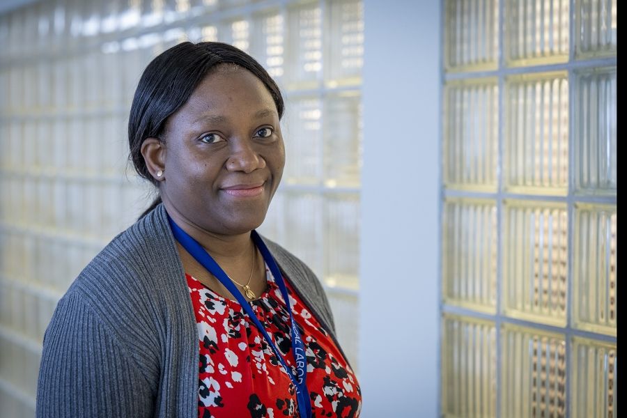 Black Staff Community Group 3 Christiana Adewale is pictured standing in a hallway at the Kingston General Hospital Site. She’s Black, has long-dark hair that’s tied up in a low ponytail and dark brown eyes. She’s wearing a bright red, white and black patterned blouse with a grey cardigan on top.