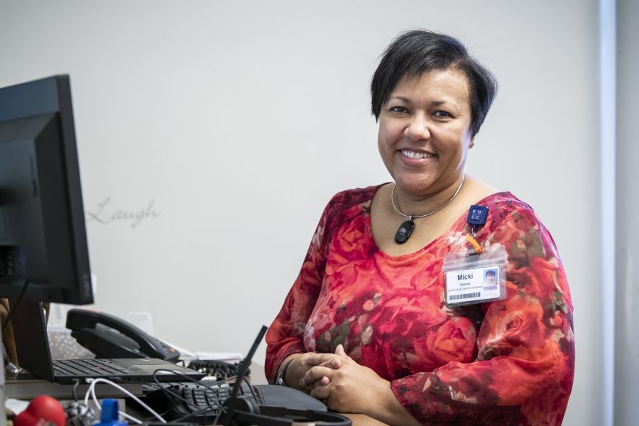 Black Staff Community Group 4 Micki Mulima is pictured sitting at her desk at the Kingston General Hospital site. She’s Black, has short-dark hair and brown eyes. She’s wearing a bright red, patterned blouse with roses.