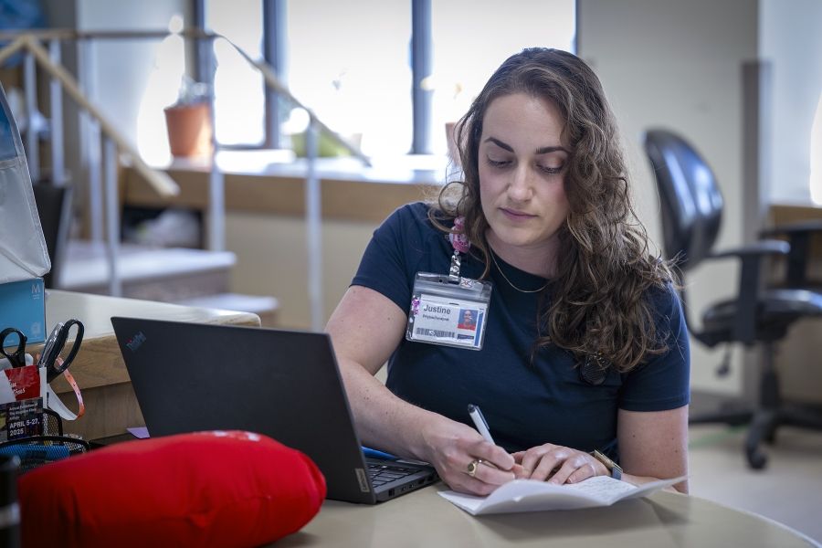 PeopleOfKHSC_Justine_White_2 Justine White is seated at a table with her computer, a cardiac surgery pillow and some paper, which she’s jotting down notes on. She has shoulder length, medium brown, wavy hair and brown eyes. She’s wearing a navy blue shirt with her KHSC ID badge.
