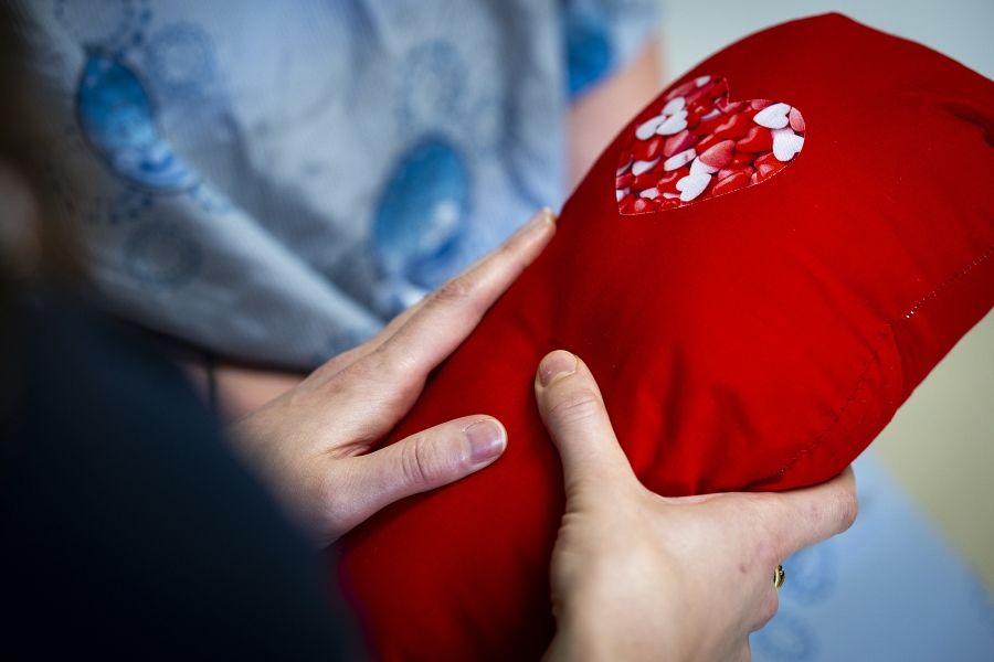 PeopleOfKHSC_Justine_White_4 A close up shot of Justine White’s hands holding a cardiac surgery pillow. The pillow is bright red with a heart near the top of the pillow.