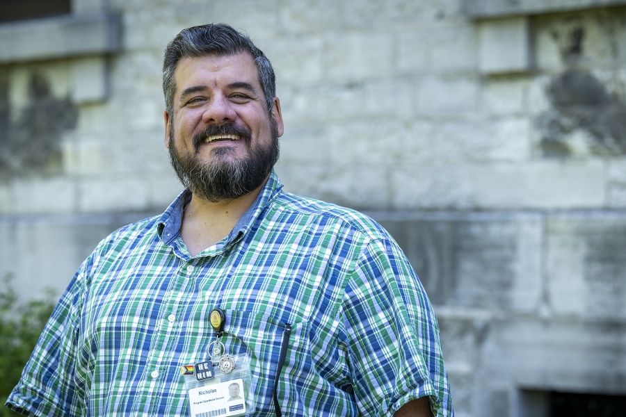 A man is standing outside Kingston General Hospital smiling directly at the camera. He has salt and pepper hair, brown eyes and a beard. He’s wearing a blue and green plaid shirt.