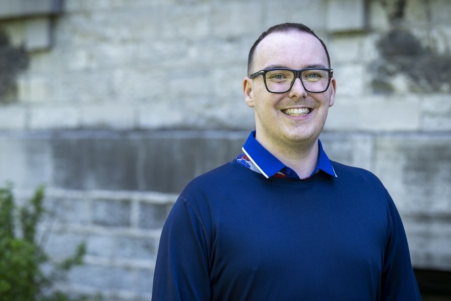 A man is standing outside Kingston General Hospital smiling directly at the camera. He has short brown hair, blue eyes and wears black framed glasses. He’s a blue sweater over a blue collared shirt.