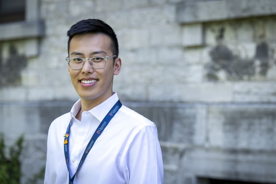 A man is standing outside Kingston General Hospital smiling directly at the camera. He has short, black hair and brown eyes. He’s wearing clear or see through framed glasses, and a white collared button down shirt.