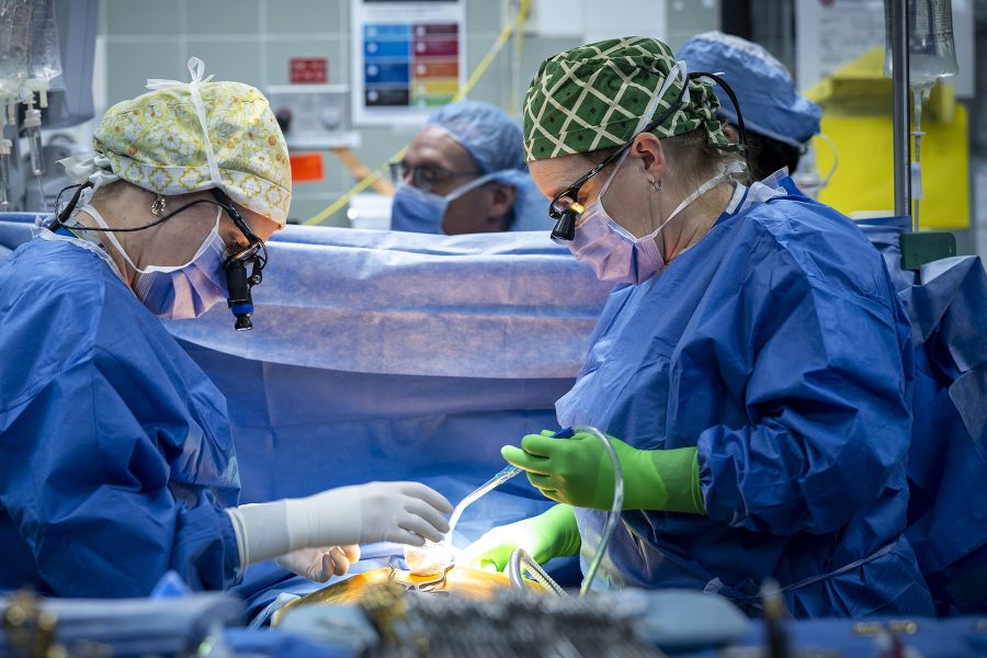 Tammy Richards is in the middle of surgery, holding instruments in her hands, while standing across from another surgeon. She’s wearing blue scrubs, gown, mask, a scrub cap and specialized glasses.