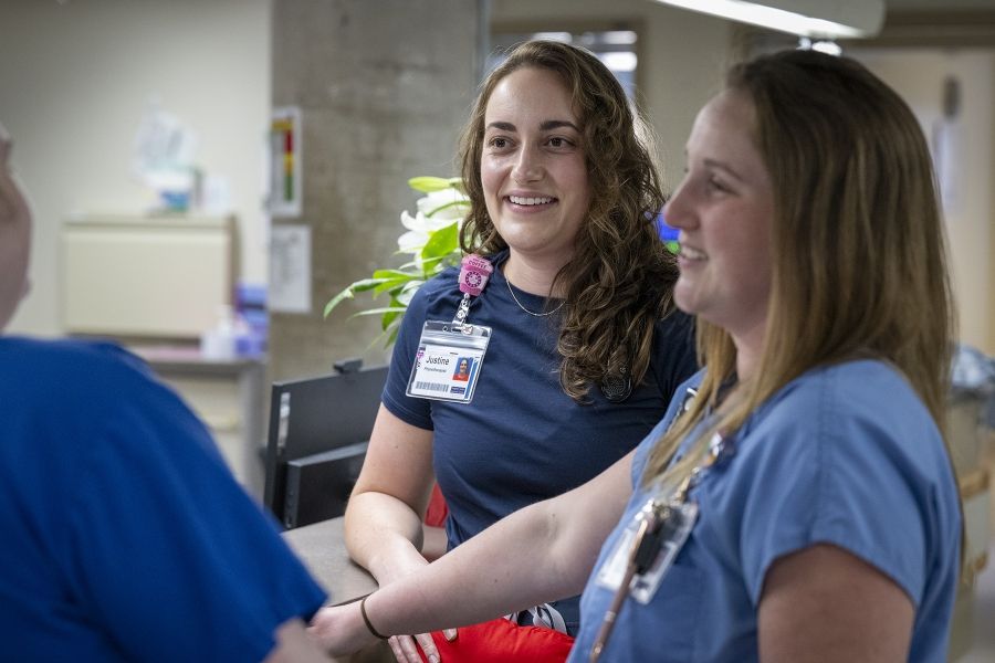 PeopleOfKHSC_Justine_White_5 Justine White is standing next to physiotherapy assistant Danielle Knapton near a care desk. The duo are seen chatting and smiling with another person whose back is to the camera.