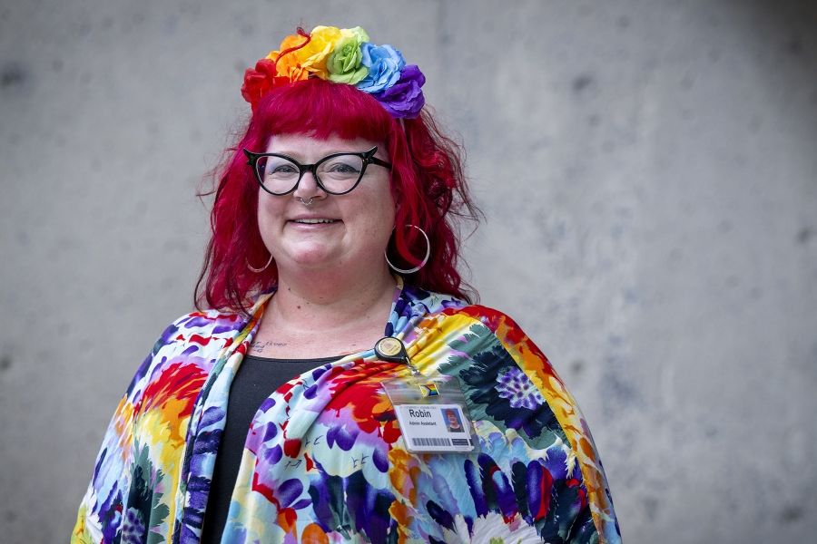 A woman is standing outside Kingston General Hospital smiling directly at the camera. She has long, bright red hair, blue eyes and is wearing black, cat eye framed glasses. She’s wearing a multi-coloured floral blouse over a black shirt, and a multi-colour flower crown on her head.