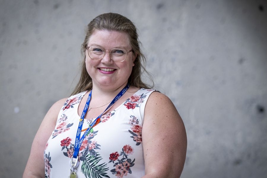 A woman is standing outside Kingston General Hospital smiling directly at the camera. She has long, straight medium brown hair, blue eyes and is wearing clear glasses. She’s a white tank top with multi-colour flowers on it.