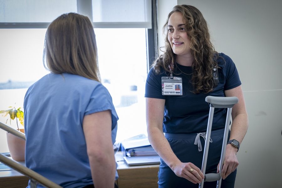 PeopleOfKHSC_Justine_White_6 Justine White is standing and putting together a crutch, while looking at physiotherapy assistant Danielle Knapton, whose back is to the camera. White has shoulder length, medium brown, wavy hair and brown eyes. She’s wearing a navy blue shirt with her KHSC ID badge.