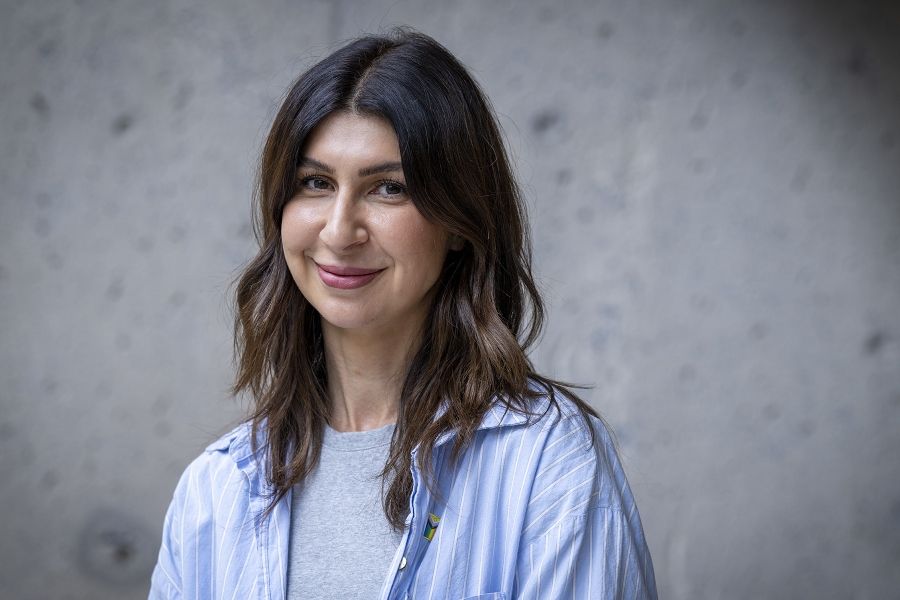 A woman is standing outside Kingston General Hospital smiling directly at the camera. She has long, wavy dark brown hair and brown eyes. She’s wearing a blue pinned stripped collared shirt with a grey t-shirt underneath.