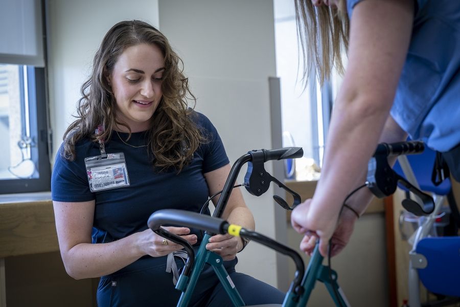 PeopleOfKHSC_Justine_White_7 Justine White is kneeling working on a walker, a mobility aid device. Physiotherapy assistant Danielle Knapton, whose back is to the camera, is holding the device. White has shoulder length, medium brown, wavy hair and brown eyes. She’s wearing a navy blue shirt with her KHSC ID badge.