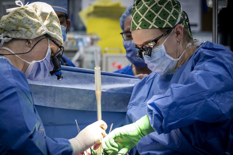 Tammy Richards is in the middle of surgery, holding instruments in her hands, while standing across from another surgeon. She’s wearing blue scrubs, gown, mask, a scrub cap and specialized glasses.