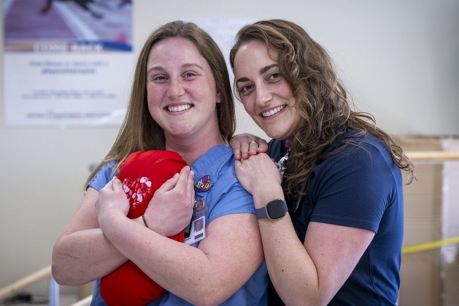 PeopleOfKHSC_Justine_White_9 Justine White and physiotherapy assistant Danielle Knapton embracing with a cardiac surgery pillow. White has shoulder length, medium brown, wavy hair and brown eyes. She’s wearing a navy blue shirt with her KHSC ID badge. Knapton has straight medium brown hair and blue eyes. She’s wearing light blue scrubs with her KHSC ID badge.