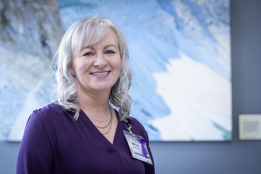 Melissa Meisner is standing in a hallway, smiling directly into the camera. Meisner has blue eyes, shoulder length, silver hair and is wearing a bright purple blouse.