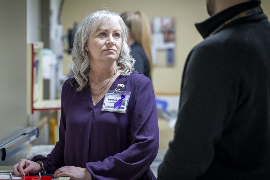 Melissa Meisner is standing next to a colleague in a hallway, looking up at them. Meisner has blue eyes, shoulder length, silver hair and is wearing a bright purple blouse.