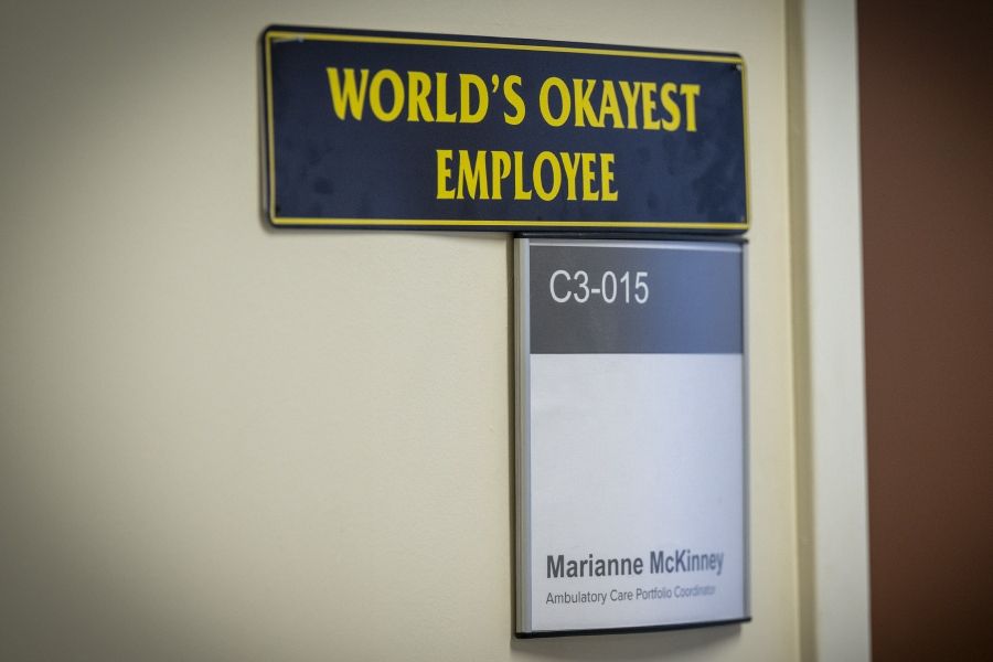 PeopleOfKHSC_Marianne_McKinney_5 A close up shot of a sign outside of Marianne McKinney’s office that says “World’s Okayest Employee.” The sign is black with bright yellow lettering.