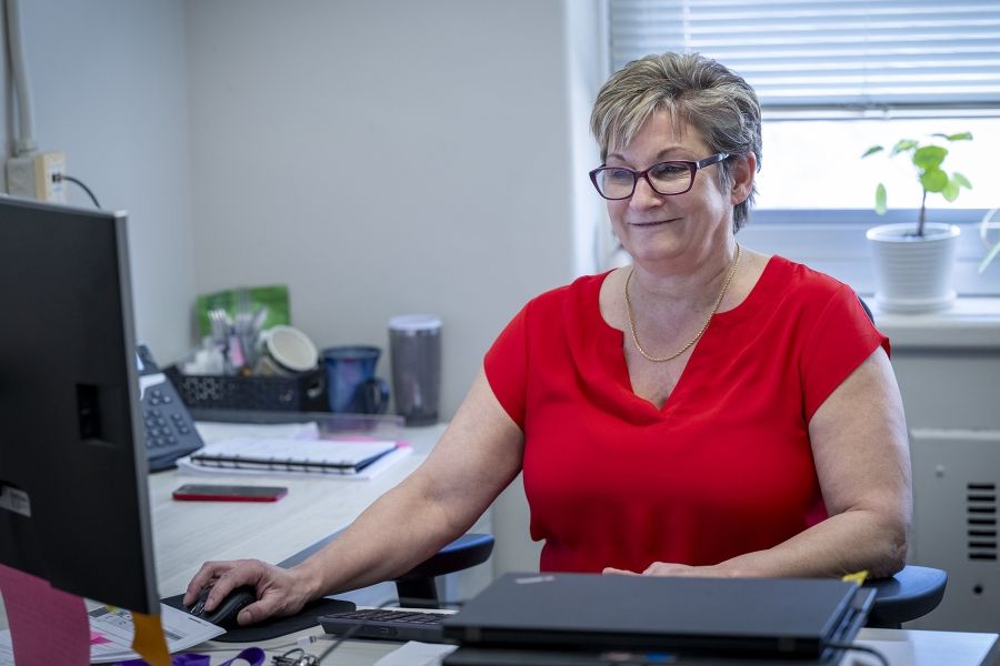 PeopleOfKHSC_Marianne_McKinney_8 A shot of Marianne McKinney sitting at her desk, smiling at her computer monitor. McKinney has short dirty blonde hair, blue eyes, wears glasses and is dressed in a bright red blouse.