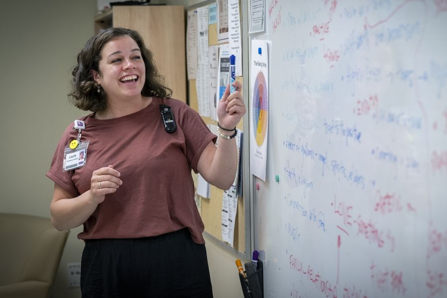 PeopleofKHSC_Laurie_Hill_2 Laurie Hill is standing in front of a white board with a dry erase marker in her hand, and smiling. She has short brown wavy hair and is wearing a dusty rose blouse and black pants.