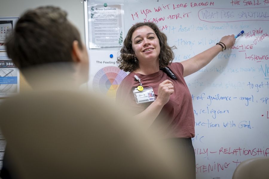 PeopleofKHSC_Laurie_Hill_3 Laurie Hill is standing in front of a white board with a dry erase marker in her hand. She’s looking out at a colleague whose back is to the camera, and smiling while pointing at something on the board with the dry erase marker. She has short brown wavy hair and is wearing a dusty rose blouse and black pants.